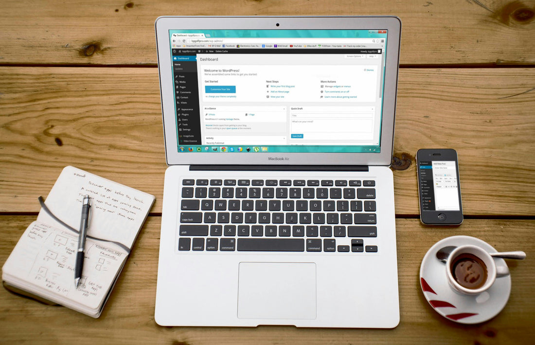 a photo of a macbook on a desk. There is a notepad on the left and a cup of coffee on the right alongside a mobile phone. On the screen of the Macbook the Wordpress backend cms is displayed.